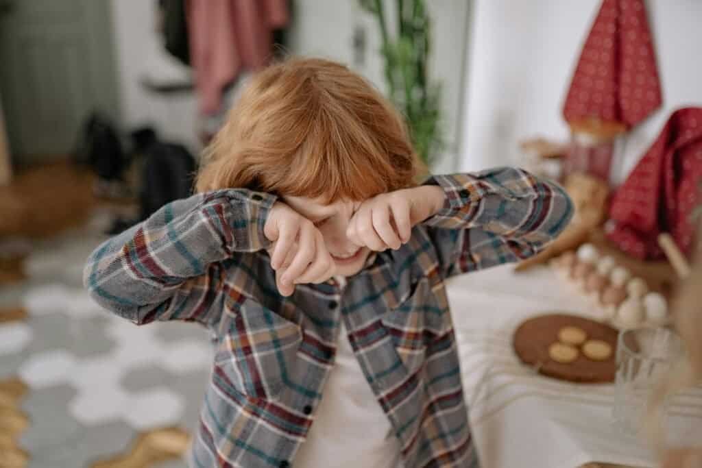 Understanding Childhood Anxiety 1 A young boy with red hair wipes his eyes in a cozy kitchen, evoking emotion and childhood innocence.