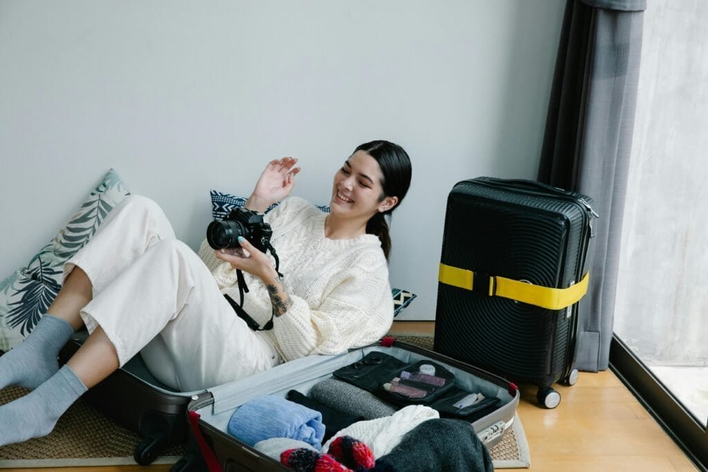 Young woman packing camera in suitcase, smiling indoors, ready for travel.